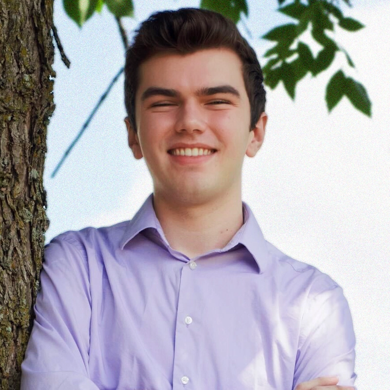 A young man with short brown hair smiles with arms crossed, leaning against a tree. He wears a light purple button-up shirt. Green leaves and blue sky are visible in the background.