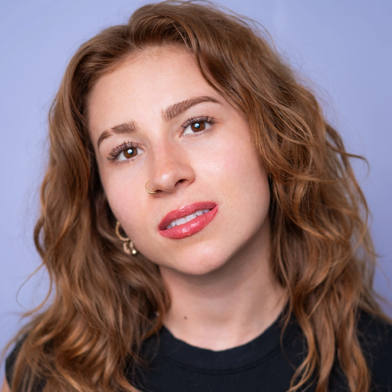 A young woman, Tessa Washburn, with wavy brown hair, wearing a black top and gold hoop earrings, looks at the camera with a neutral expression. She has a nose ring, subtle makeup, and glossy lips. The background is a soft, light purple color.