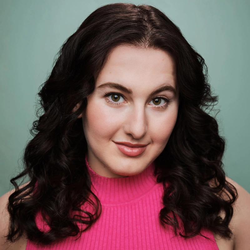 Olivia Wasser, a woman with wavy dark hair, wears a bright pink sleeveless knit top and smiles softly against a plain teal background. Her makeup is natural, and she looks directly at the camera, conveying confidence and warmth.