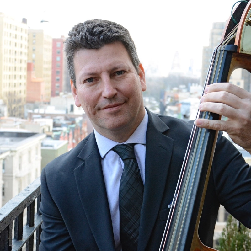 A man in a suit and tie holds an upright bass on a balcony overlooking a cityscape with tall buildings. He smiles slightly at the camera. The scene appears to be taken during the day with a clear, bright sky in the background.