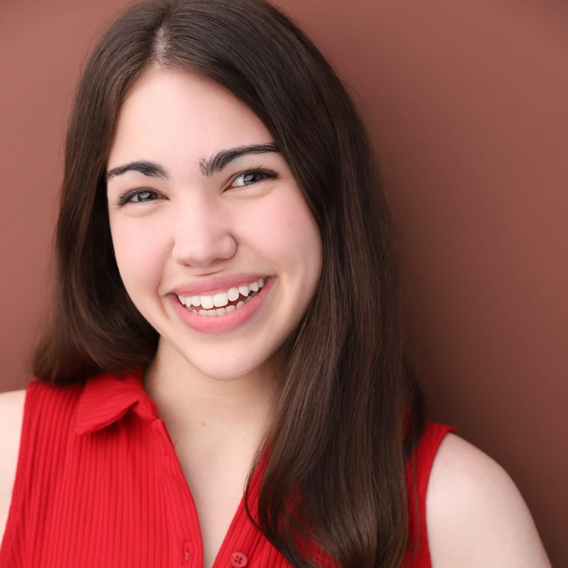 A young woman with long brown hair smiles at the camera. She wears a sleeveless red shirt and stands against a reddish-brown background. Her teeth show as she smiles, and her hair is parted in the middle, cascading over her shoulders, capturing Jenna Weisbach’s warm and approachable demeanor.