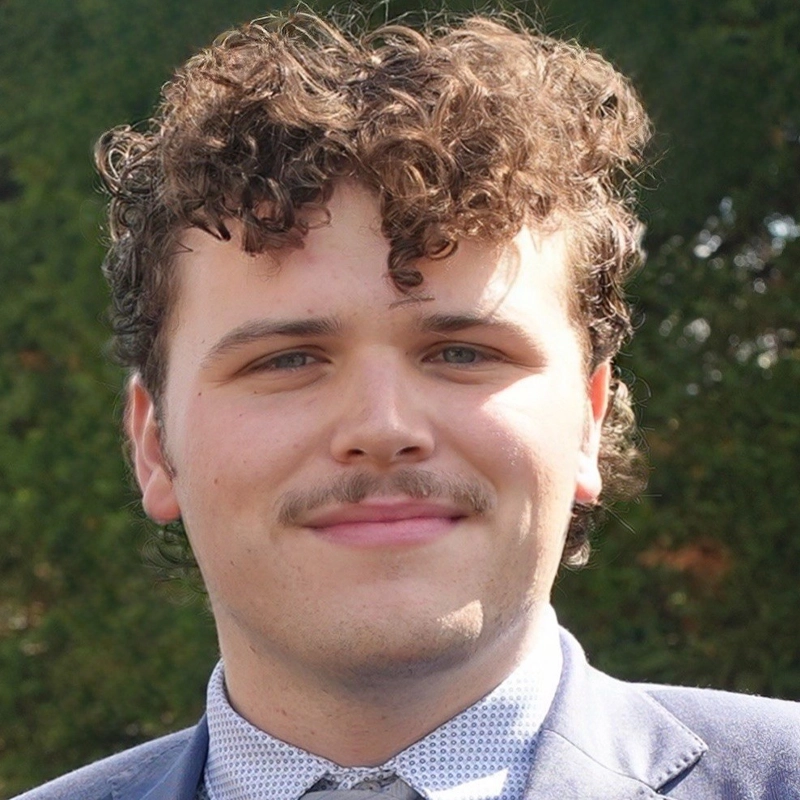 Aidan C. White, a young man with curly brown hair and a mustache, smiles at the camera. He wears a blue patterned dress shirt, a gray tie, and a blue jacket. The background shows blurred green foliage in sunlight.