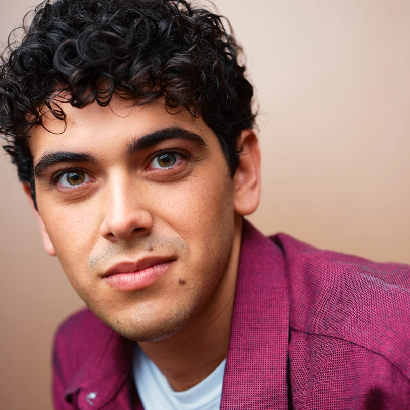 A young man with curly dark hair and green eyes looks into the camera. Oliver Whitehouse has a small mole on his cheek, wears a maroon jacket over a white shirt, and is posed against a plain beige background.