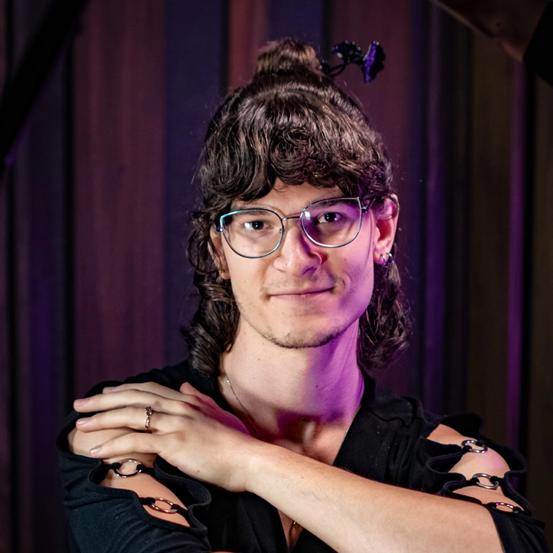 A young person with wavy brown hair in a half-up style, wearing glasses and a black shirt with cutout sleeves, crosses their arms. The background features vertical wooden panels with purple lighting, adding an atmospheric touch reminiscent of Willow Wakar’s signature style.