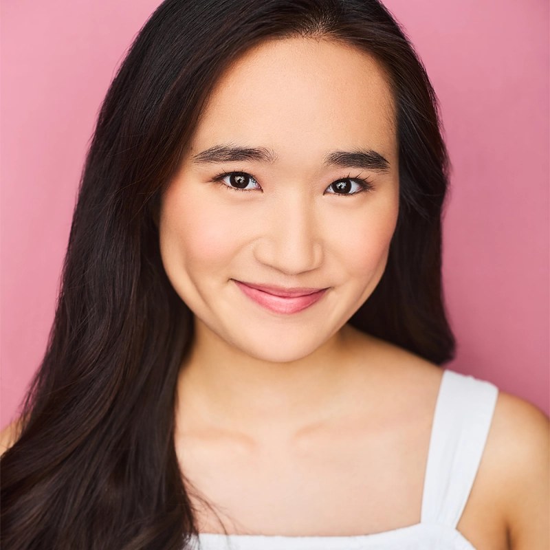 A young woman with long dark hair smiles gently, showing dimples. She has smooth skin, natural makeup, and wears a sleeveless white top. The background is soft pink, creating a cheerful and warm atmosphere.