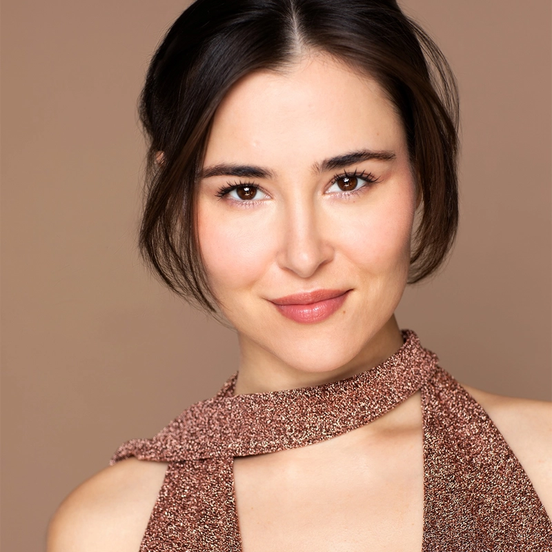 A woman with fair skin and dark brown hair, parted in the middle and tucked behind her ears, smiles softly. Reminiscent of Kseniya Yumasheva’s understated elegance, she wears a sparkly, textured, rose-gold halter top with a matching neck detail. The background is a solid, warm beige color.