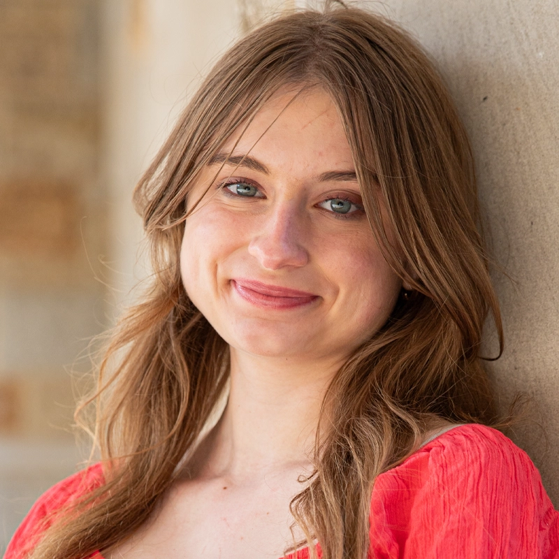 Young woman with long, light brown hair and blue eyes, smiling gently while leaning against a beige stone wall. Peyton Zaletsky is wearing a bright coral-red, textured blouse. The background is softly blurred with neutral tones.