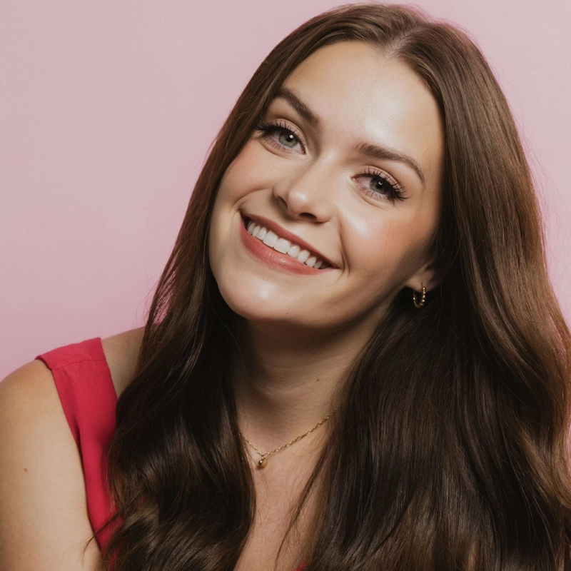 A young woman with long brown hair smiles brightly at the camera. Jacquelyn vonAschwegue wears a pink sleeveless top, gold earrings, and a delicate gold necklace. The background is solid light pink, creating a cheerful and warm atmosphere.