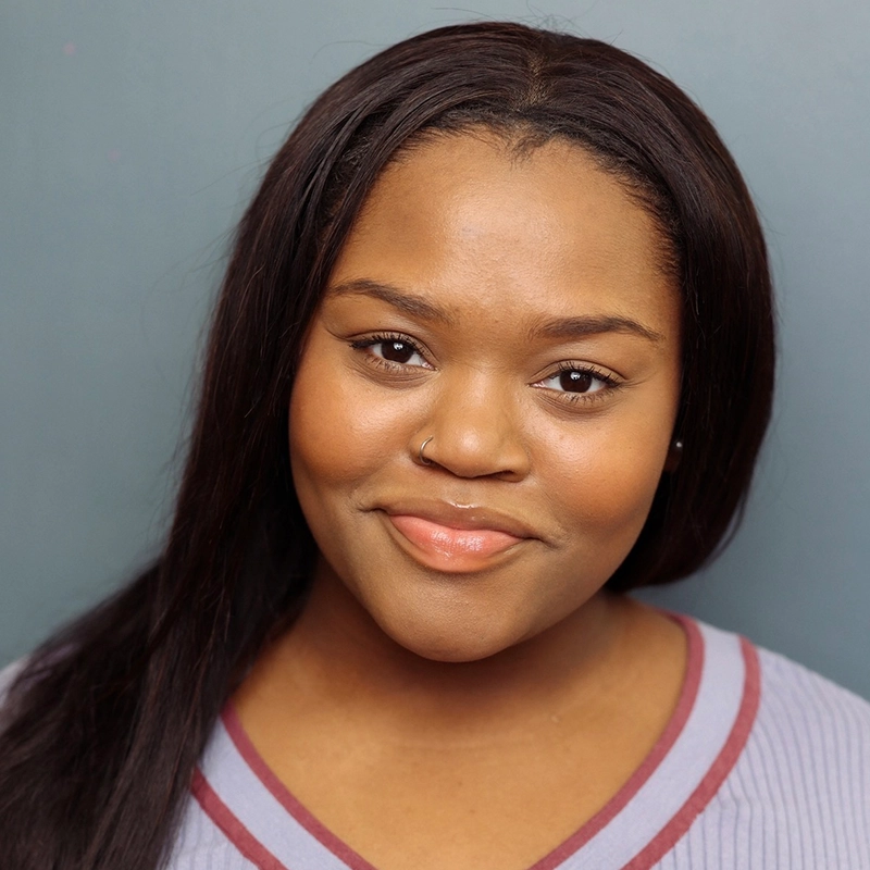 A young woman with long, straight dark hair smiles gently. She has a nose ring, warm brown skin, and wears a purple top with a grey trim. The background is a plain, muted blue. This is Leah Mossman.