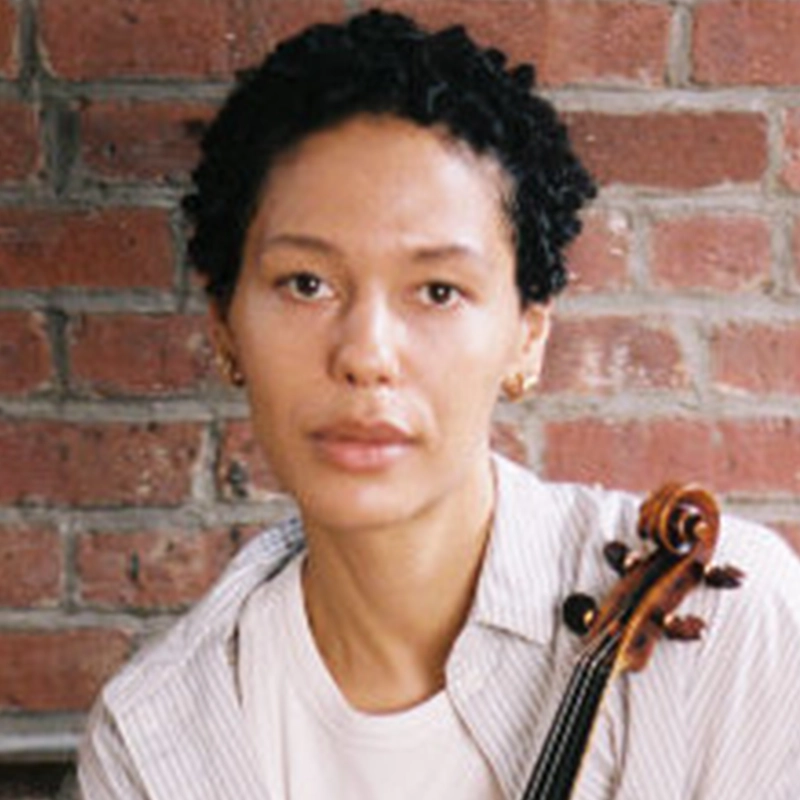 Alexandria Hill, a person with short, curly hair, looks directly at the camera. They wear a light-colored collared shirt and hold the neck of a violin over their shoulder against a brick wall background.