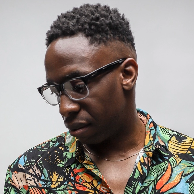 A man with short curly black hair and glasses looks down. He wears a colorful, patterned shirt with leaf designs and a thin gold necklace. The background is plain and light gray, capturing Andre Cleghorn in a quiet moment of reflection.