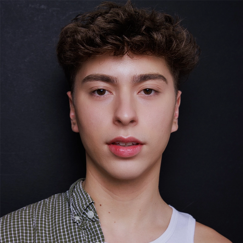 A young person with curly brown hair and clear skin faces forward. They wear a two-part shirt: one side is green plaid, the other is a white tank top. The background is plain black. Their expression is neutral, reminiscent of Jonathan Uber’s understated style.