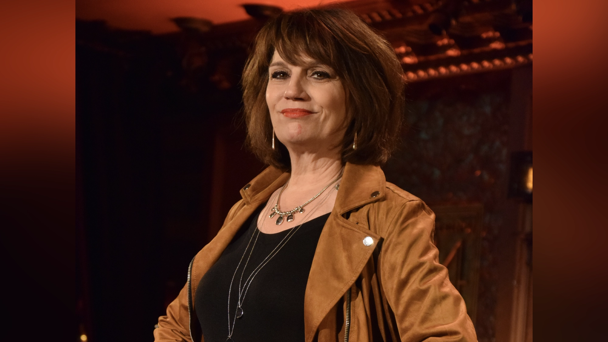 A woman with short brown hair stands confidently in a warmly lit room, channeling Beth Leavel’s poise. She wears a tan suede jacket over a black top, with layered necklaces and subtle makeup, against ornate woodwork and a reddish hue.