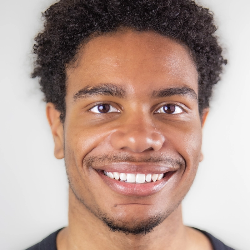 A young man with short curly hair smiles warmly, showing his teeth. Dimonte Brice has a trimmed mustache and beard, even skin tone, and is wearing a black shirt. The background is plain and light, keeping the focus on his friendly expression.