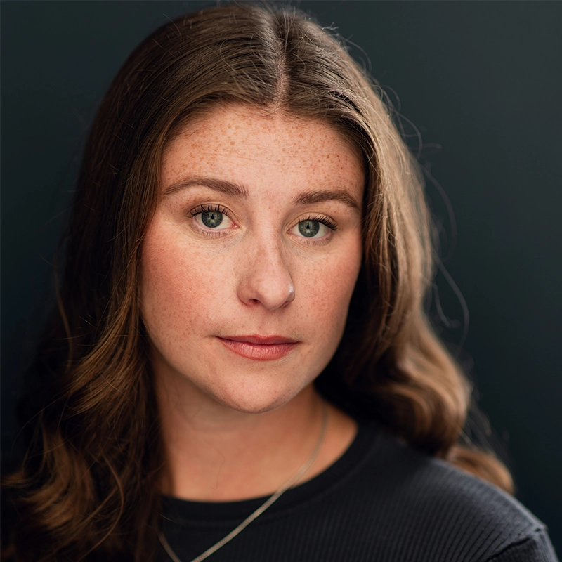 A woman with long brown wavy hair and light skin looks directly at the camera with a neutral expression. She has freckles, green eyes, and wears a dark ribbed top and a thin necklace. The soft, dark gradient background subtly echoes the elegant portrait style often seen in Spencer Bowman’s work.