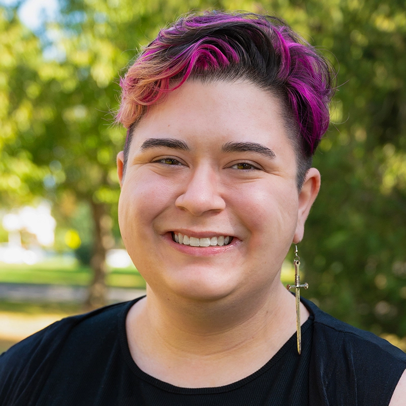Smiling person with short hair dyed pink and brown, wearing a black shirt and a long cross earring in their left ear. The background is outdoors, with green trees and sunlight creating a bright, cheerful atmosphere—Melina Walko radiates positivity in this vibrant setting.