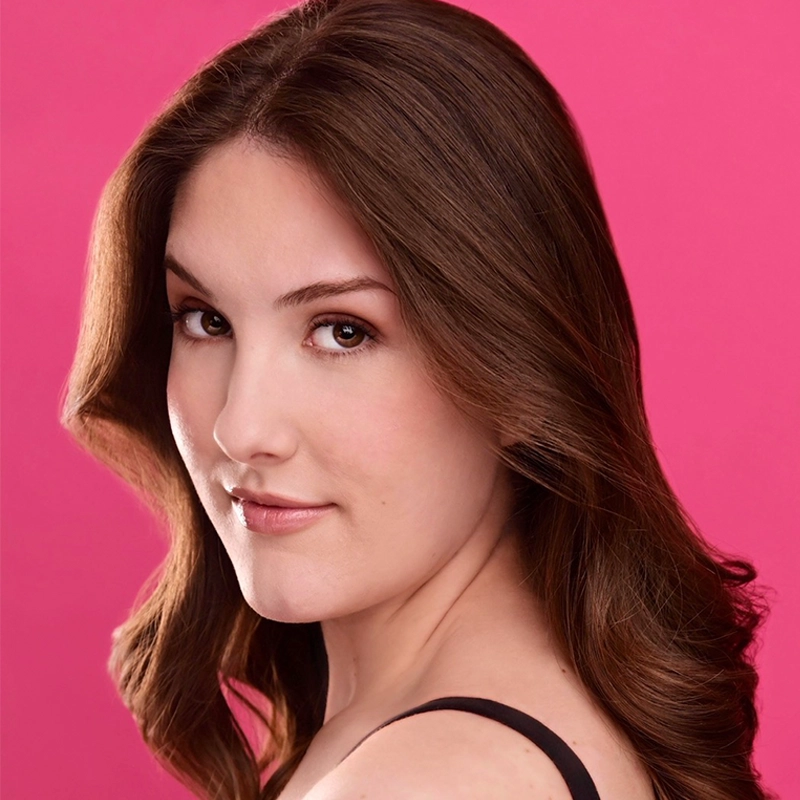 A young woman with long, wavy brown hair poses in front of a bright pink background. She wears a black top and has natural makeup with soft pink lips, gazing confidently over her shoulder toward the camera in this signature Jonathan Uber portrait style.