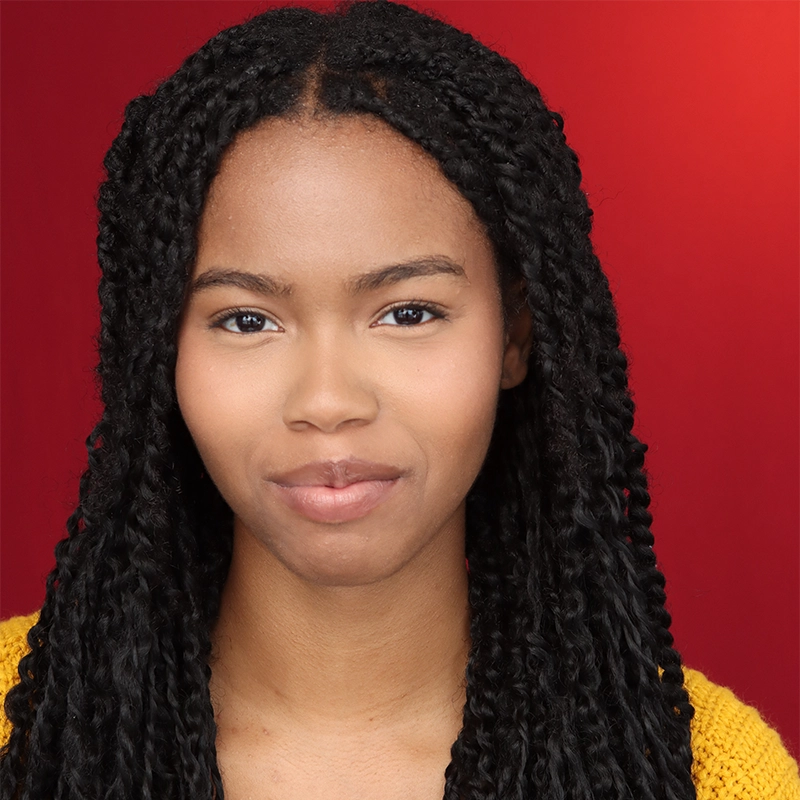 A young woman with long, dark braided hair wears a yellow sweater and smiles softly. Rayana D. Briggs faces forward against a solid red background. Her expression is calm and confident, and her skin tone is medium brown. The image is well-lit and clear.