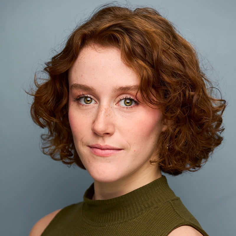 Phoebe Kleehammer, a young woman with fair skin, short curly red hair, and green eyes, looks into the camera with a slight smile. She wears a sleeveless olive-green top against a plain light blue background. The lighting is soft, and her makeup is natural.