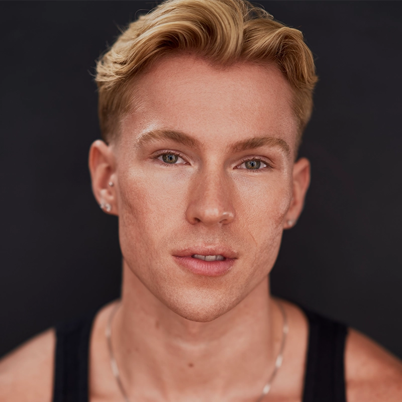 A young man with short, styled blonde hair and light skin gazes forward with a neutral expression. He wears a black tank top, small earrings in both ears, and a thin silver necklace—a look that reflects the understated confidence of Jonathan Uber. The background is dark and out of focus.
