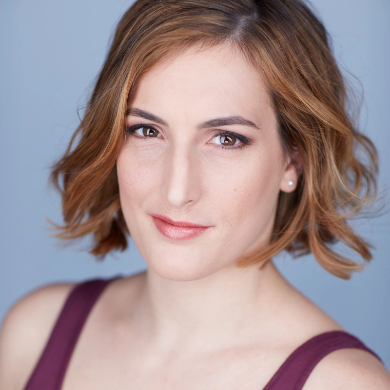 Samantha Chiodo, a woman with fair skin and wavy, chin-length light brown hair, smiles slightly. She wears natural makeup, stud earrings, and a sleeveless maroon top. The background is a soft, solid light blue, and the lighting is even, emphasizing her facial features.