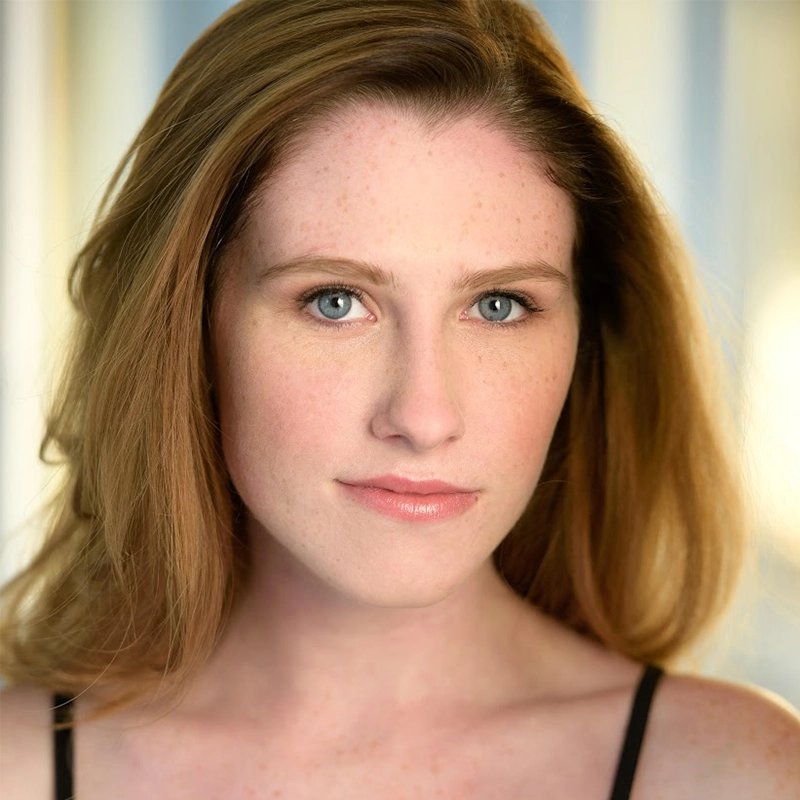 A young woman with long, straight, light brown hair and blue eyes looks directly at the camera. She has fair skin with freckles, light makeup, and a neutral expression. Melina Walko is wearing a black top. The background is softly blurred with blue and yellow tones.