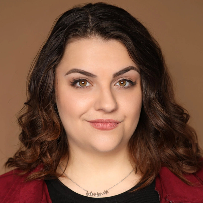 A woman with wavy brown hair and green eyes smiles slightly. Christina Thompson wears natural makeup, a maroon jacket, a black top, and a pendant necklace. The solid light brown background enhances her confident, approachable expression.