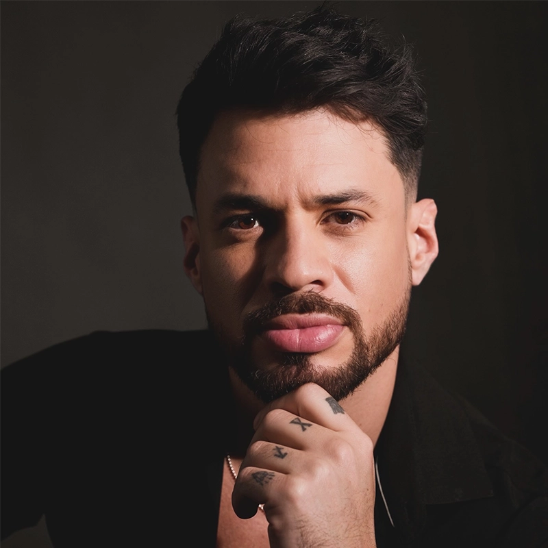 A man with short dark hair, a beard, and a mustache looks directly at the camera. He rests his chin on his hand, displaying tattoos on his fingers. Claudio Medina Jr. wears a black shirt and sits against a dark, neutral background.