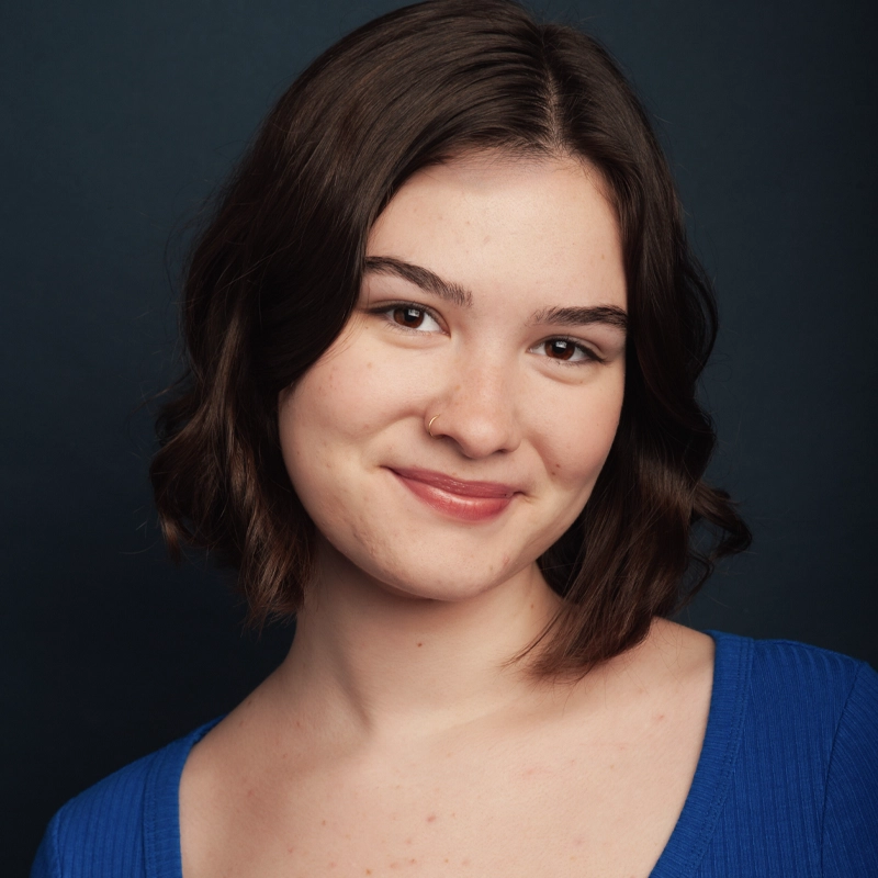 A young woman named Lilly with short brown hair smiles gently at the camera. She wears a blue top and has a nose ring. The background is dark, emphasizing her face. Her head is slightly tilted, and her expression is relaxed and friendly.