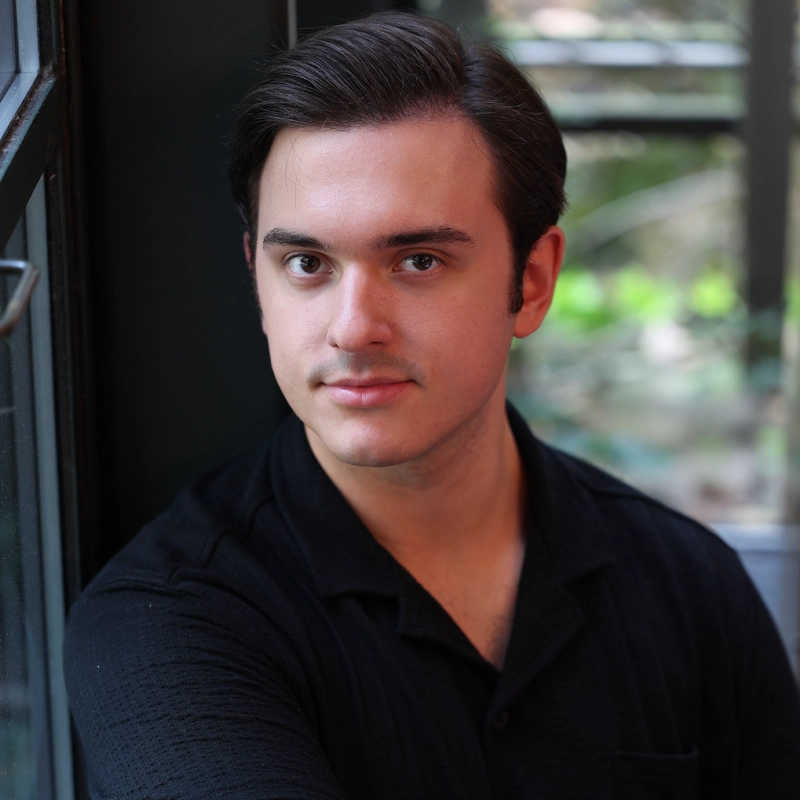 A young man with fair skin and dark hair, swept back, sits indoors by a window. He wears a black collared shirt and looks directly at the camera with a slight smile. The softly blurred background reveals some greenery outside, creating a calm setting around Graham Douglas.