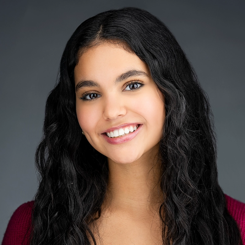 Daniela Vicente, a young woman with long, dark, wavy hair, smiles at the camera while wearing a maroon top against a solid grey background.