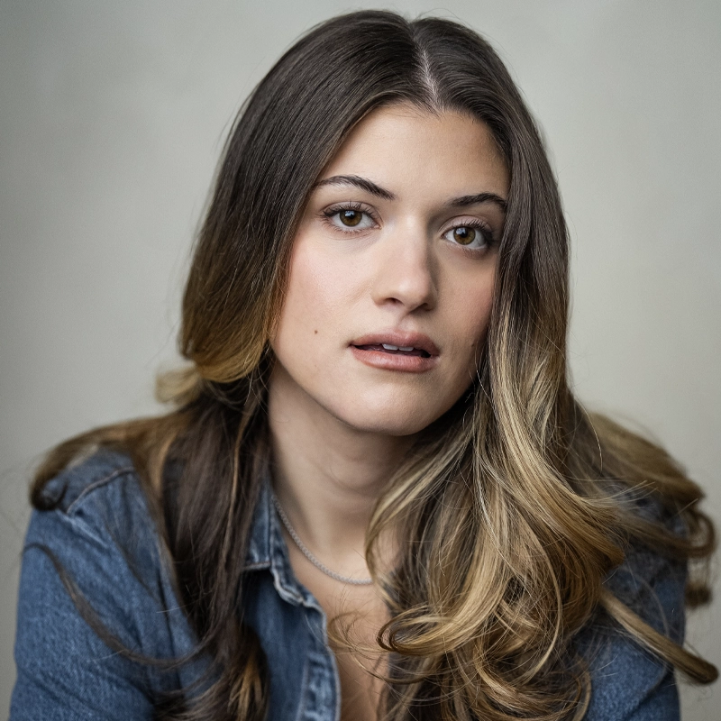 A young woman with long, wavy brown hair and lighter highlights gazes softly at the camera. Eve Dillingham wears a denim shirt over a light top and a thin necklace. The background is neutral and out of focus, drawing attention to her calm, expressive face.