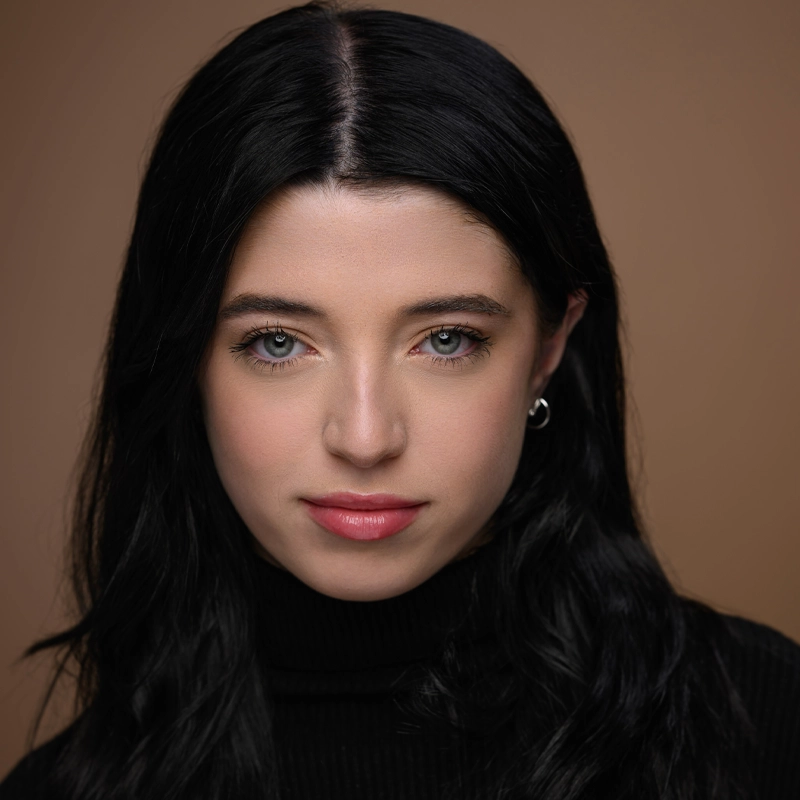 A woman with long black hair, light skin, and green eyes looks directly at the camera, wearing a black turtleneck sweater and small silver hoop earrings. The background is a plain light brown color. She has a neutral, slight smile expression.