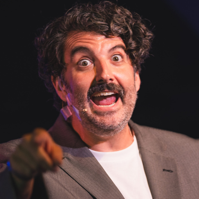 A man with curly dark hair and a mustache, wearing a gray blazer and white shirt, speaks animatedly on stage with his mouth open and eyes wide. He wears a headset microphone and points toward the camera, with a blurred dark background.