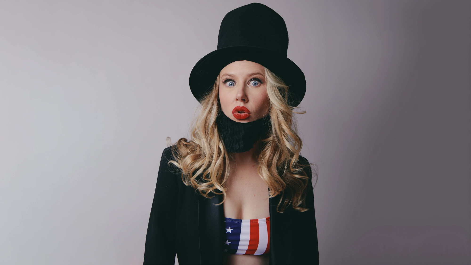 A woman with long blonde curls wears a black top hat, a fake black beard, a black jacket, and a bandeau top with the American flag design. She looks surprised, with wide eyes and red lips, against a plain light background, channeling an eccentric vibe reminiscent of Ellie MacPherson’s bold style.