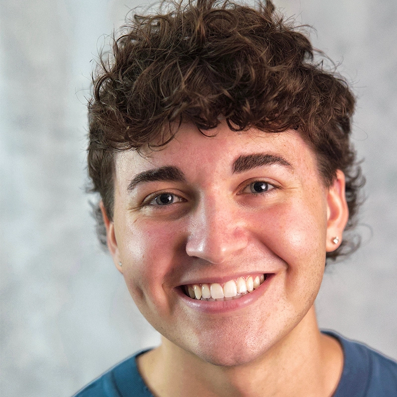 A young person with curly brown hair smiles warmly at the camera. Kinsley Stephens has light skin, dimples, and wears small earrings in both ears. The background is softly blurred in neutral tones, and they are wearing a blue shirt.