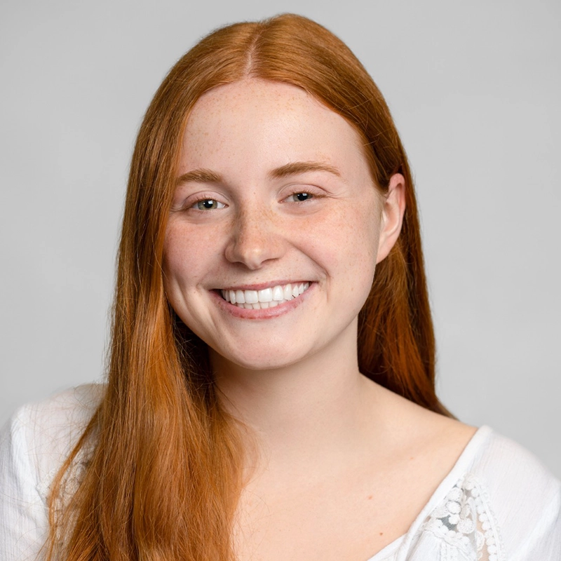 A young woman with long red hair smiles at the camera. She has fair skin, light freckles, and is wearing a white blouse. The background is plain and light gray.