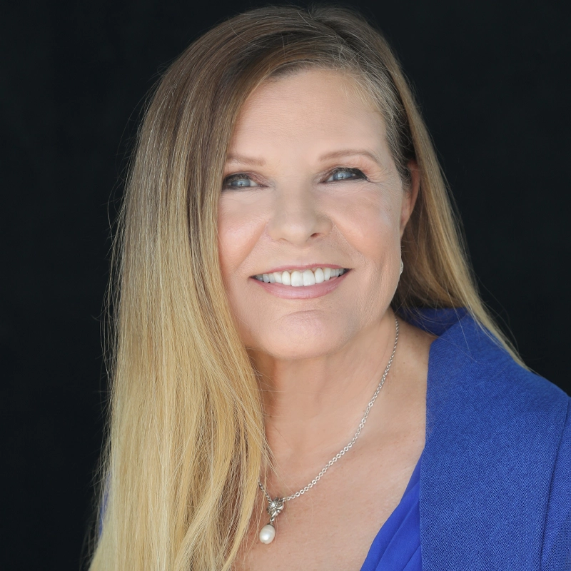 Dianne Fraser, a woman with long, straight blonde hair, smiles at the camera. She wears a bright blue blazer over a blue top, a silver necklace with a pearl pendant, and has a natural makeup look. The background is solid black.
