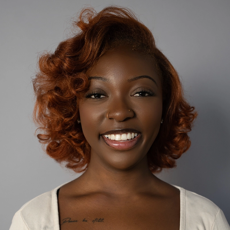 A smiling woman with medium-brown skin and curly, shoulder-length auburn hair stands against a plain gray background. She wears a light-colored top and has a small nose ring and script tattoo on her right collarbone, reminiscent of the portrait style favored by Spencer Bowman.