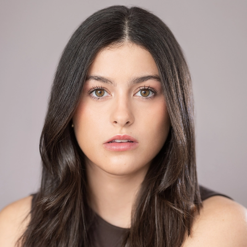 Gabriela Descartes López, a young woman with long, straight, dark brown hair and light green eyes, looks directly at the camera with a neutral expression. She has fair skin, natural makeup, and wears a sleeveless dark top against a plain, softly-lit gray background.