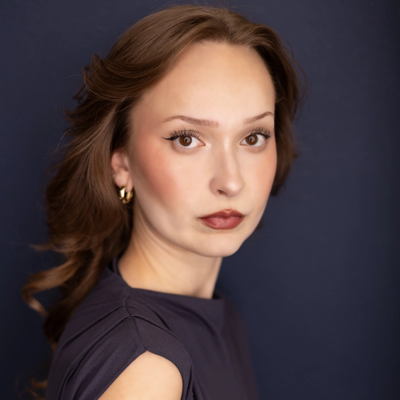 Emma Gardner, a woman with wavy brown hair, wearing gold hoop earrings and a navy top, poses against a dark blue background. She gazes at the camera with a neutral expression, and has natural makeup with defined eyebrows and soft red lipstick.
