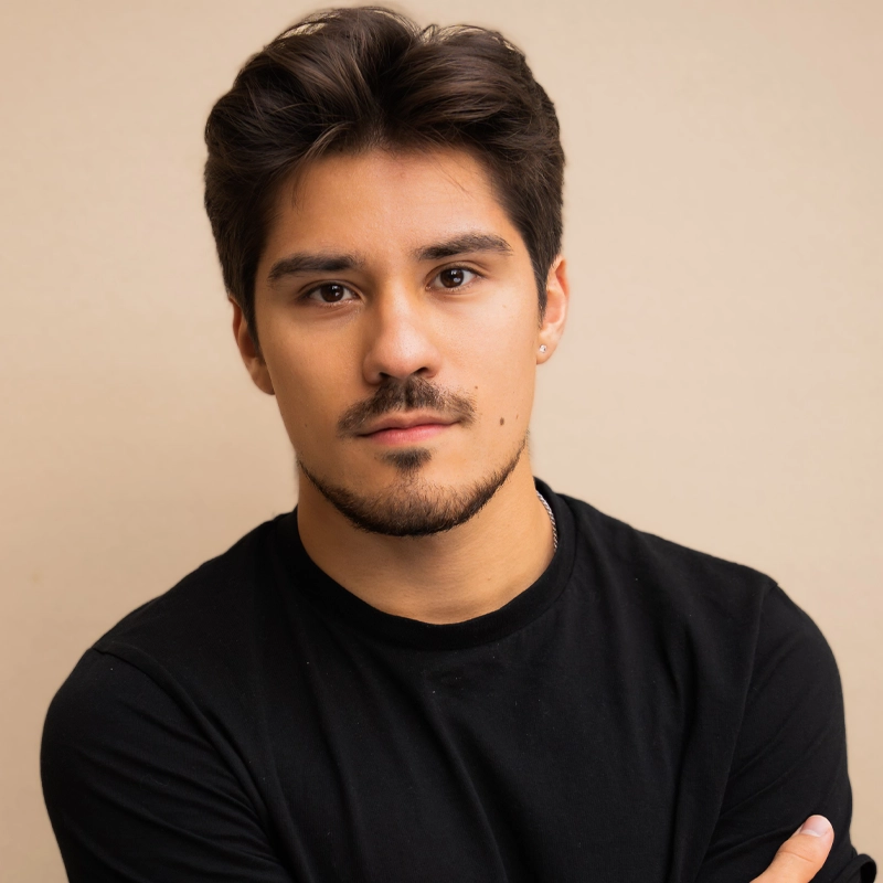 A young man with short, dark hair and facial hair, identified as Leo Gonzalez, looks at the camera. He wears a black shirt and has a neutral expression. The plain light beige background highlights his crossed arms. Image is well-lit and clear.
