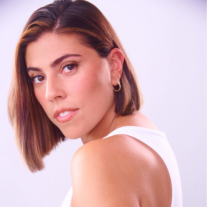 A woman with straight, shoulder-length brown hair looks over her shoulder. She wears a white sleeveless top and gold hoop earrings. Her expression is calm, and the background is a soft light purple gradient, subtly highlighting Sarah Hamaty’s poised presence.