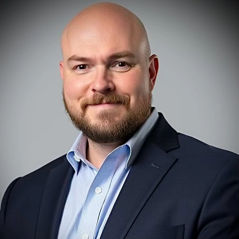 A bald man with a beard, wearing a navy blue suit jacket over a light blue collared shirt, smiles softly at the camera. The background is a simple gray gradient, giving the portrait a professional and formal appearance.