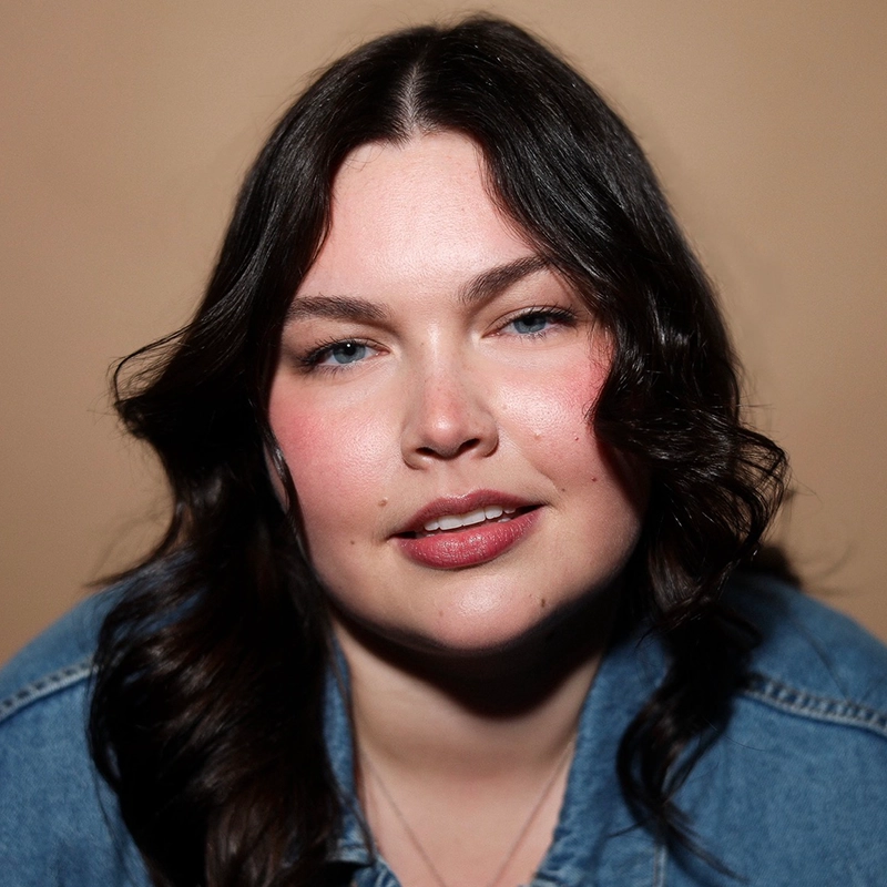 Phoebe Kleehammer, a woman with wavy dark brown hair and blue eyes, smiles softly. She wears a denim jacket and light makeup with pink blush. The background is plain and beige, and the lighting highlights her face. Her expression appears relaxed and confident.