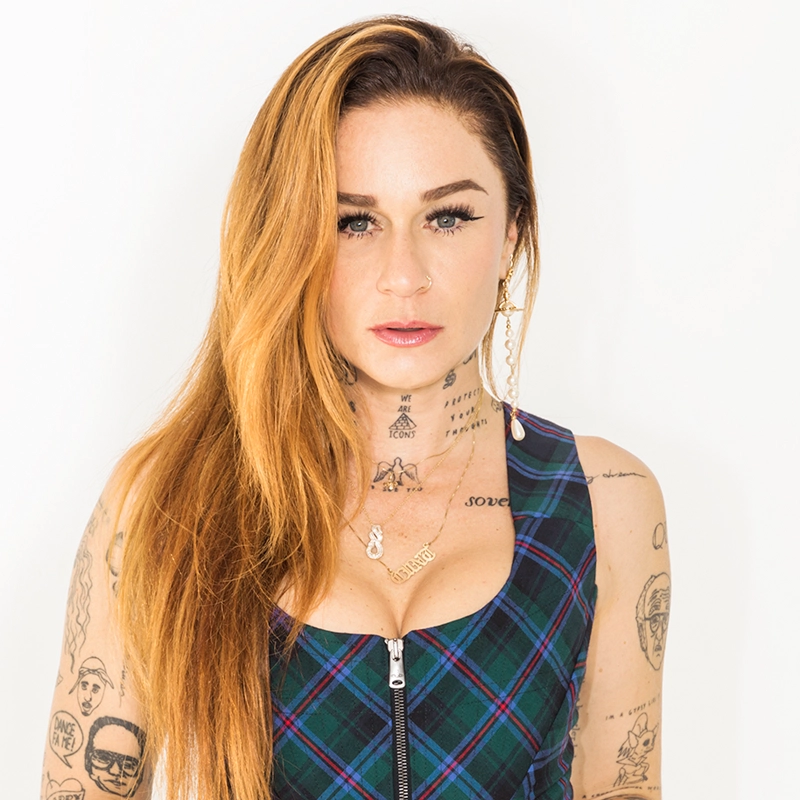 Sarah Hudson has long, reddish-blonde hair and numerous neck and arm tattoos as she looks directly at the camera. She wears a dark green and blue plaid sleeveless top with a front zipper and gold pendant necklaces against a plain, bright white background.