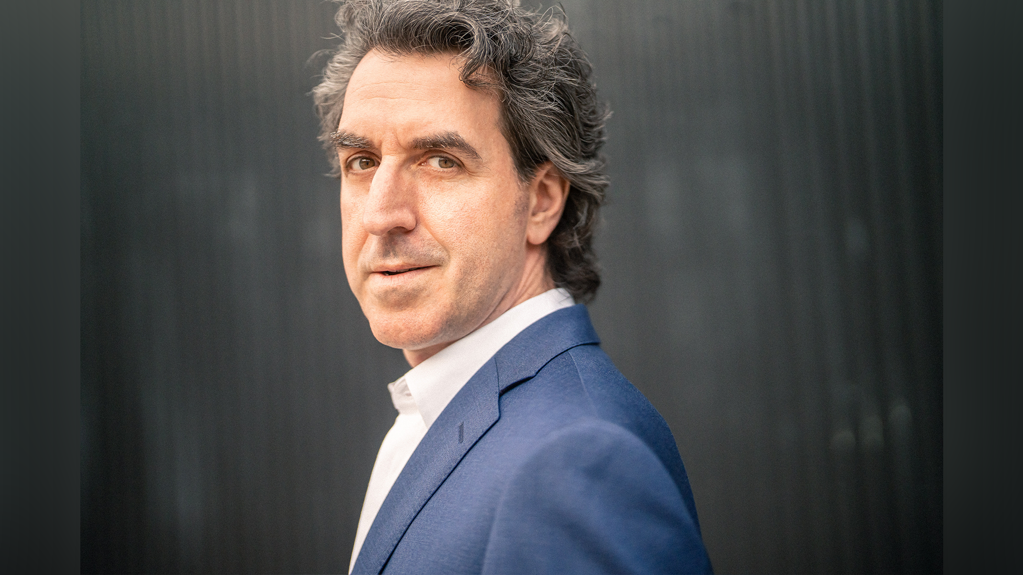 A man with wavy gray hair, wearing a blue suit jacket and white shirt, stands outdoors in profile but looks toward the camera. The background is blurred and dark, making him the central focus of the image.