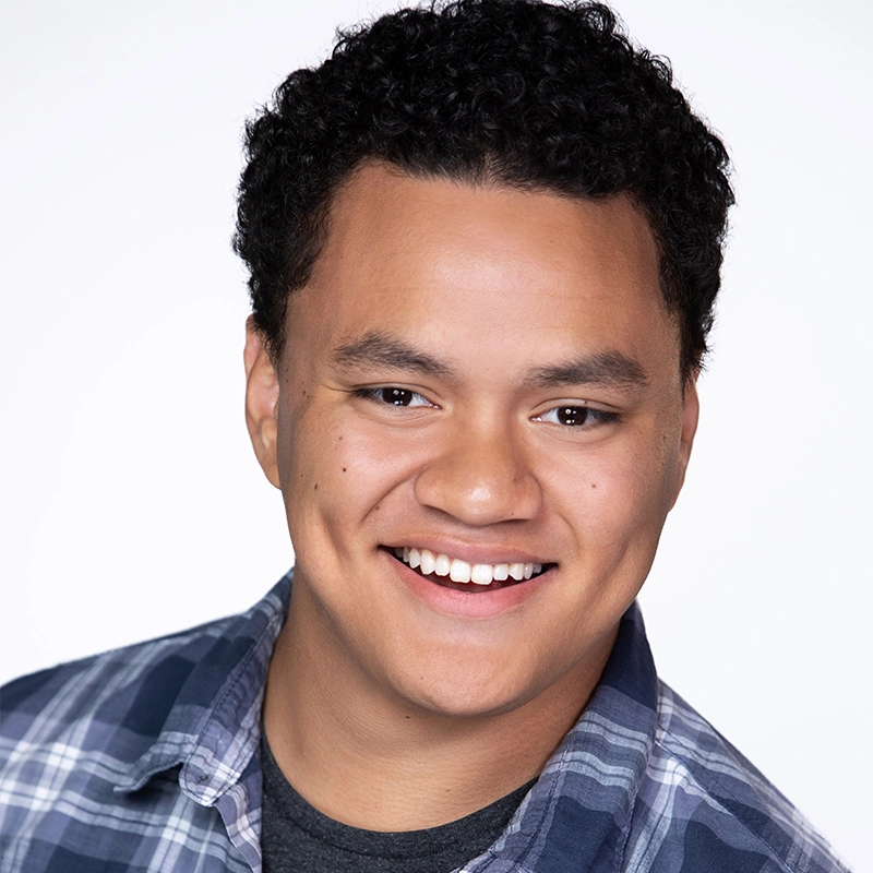 A young man with short, curly black hair smiles at the camera. He wears a blue plaid shirt over a dark gray t-shirt. The background is plain white, and the lighting is bright and even. Spencer Bowman’s expression is friendly and approachable.