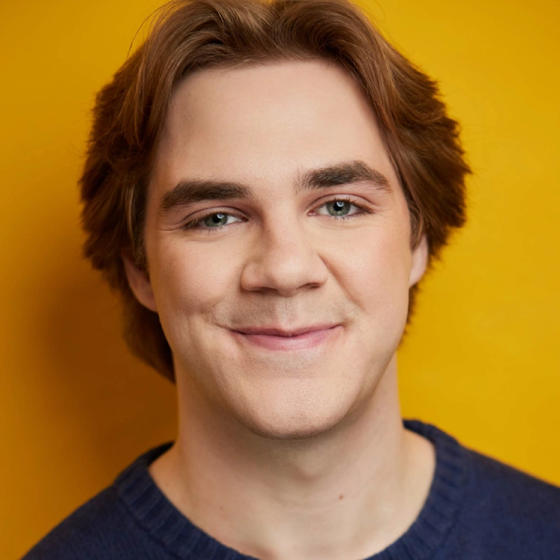 A young person with fair skin, light brown hair, and blue eyes smiles at the camera. Leah Mossman wears a dark blue sweater and is posed against a bright yellow background. The lighting is soft and even, creating a warm, friendly atmosphere.