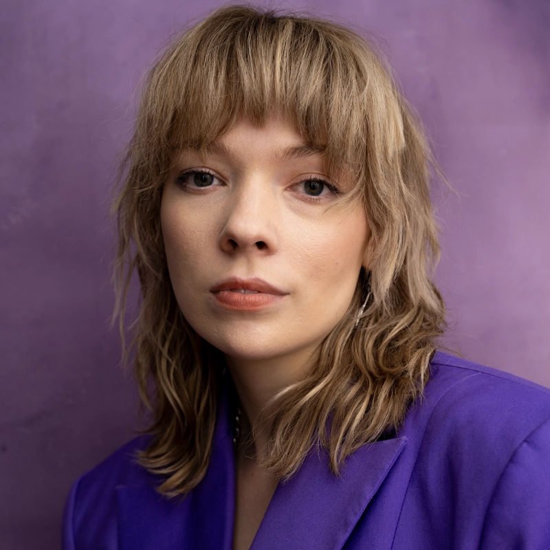 Caroline Jewell, a woman with light wavy hair and bangs, wears a purple blazer, looking into the camera with a neutral expression. The softly lit background is a muted purple shade, complementing her outfit. Her makeup is natural and understated.